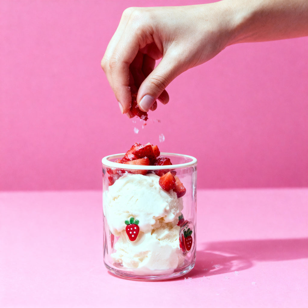 Hand sprinkling red berries onto a glass of ice cream with strawberry design on a pink background
